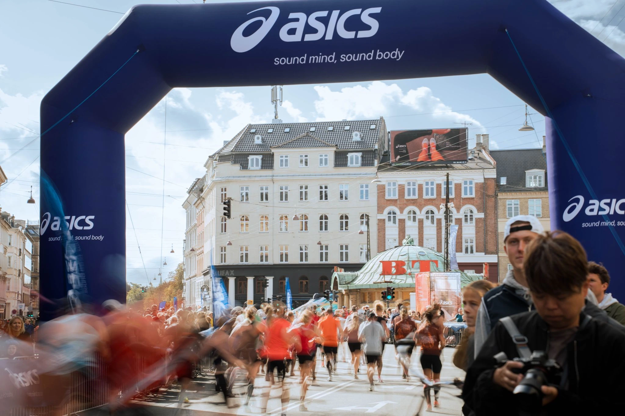 Corredores atravesando un arco de ASICS en Copenhague mientras un billboard de la marca aparece visible al fondo de la ruta.
