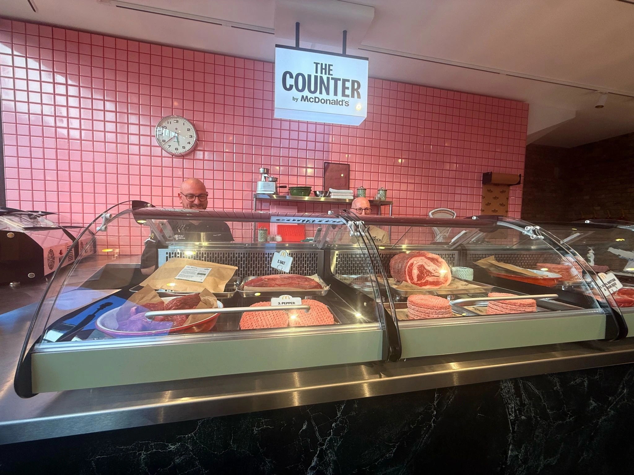 Interior de The Counter by McDonald’s con vitrinas de carne, piezas de res y señalización de los ingredientes en un espacio de azulejos rosas.